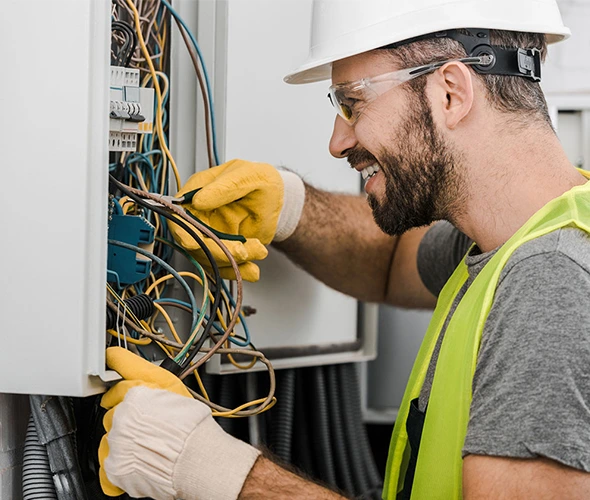 Smiling electrician in yellow safety gloves repairing electrical wires inside a junction box.